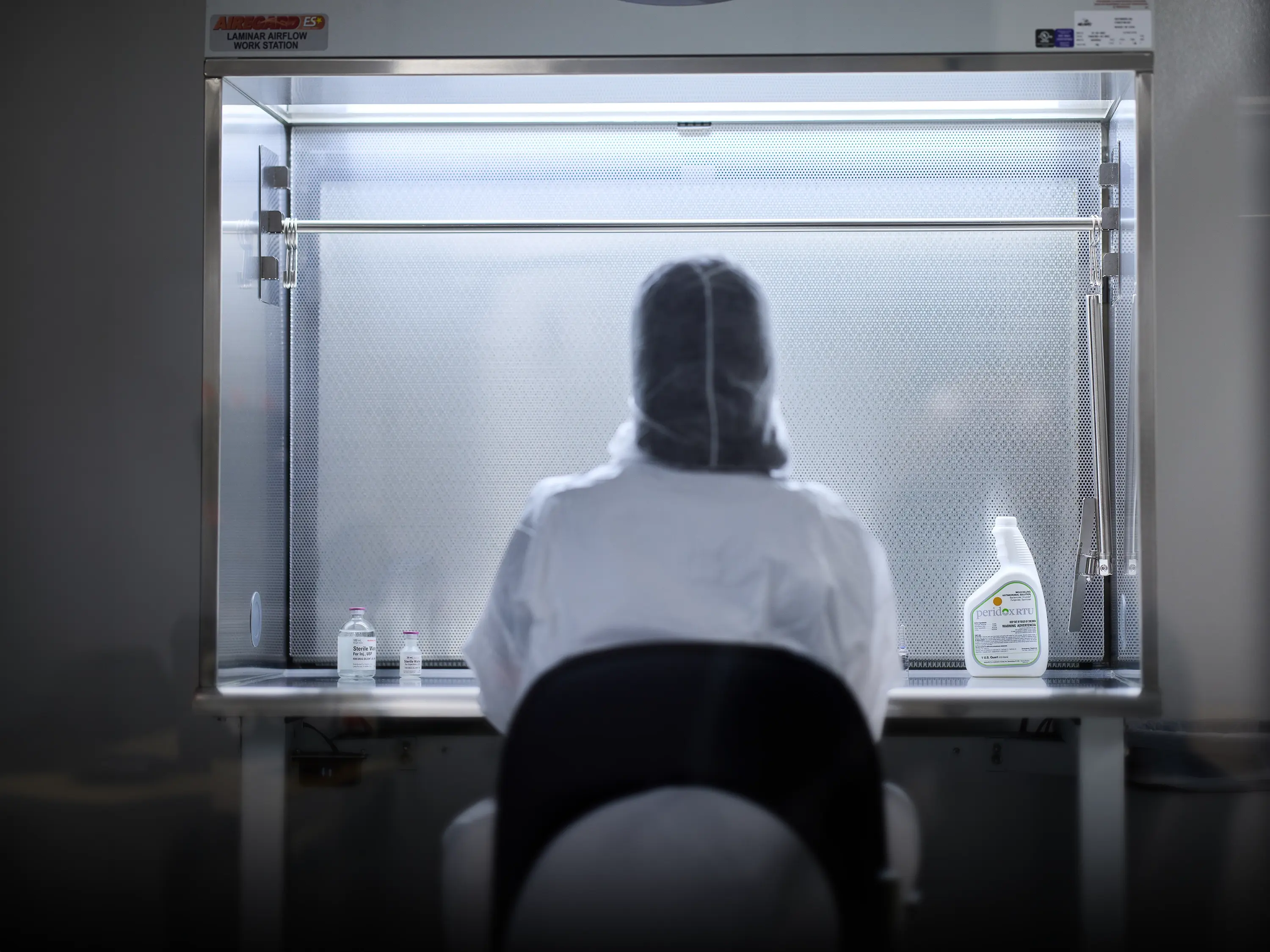 Operator in protective cleanroom garments seated at a laminar airflow workstation with supplies arranged on the work surface.